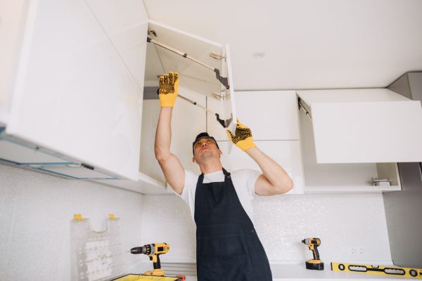 Maintenance man installing kitchen furniture in a black apron and yellow gloves using a power skrewdriver